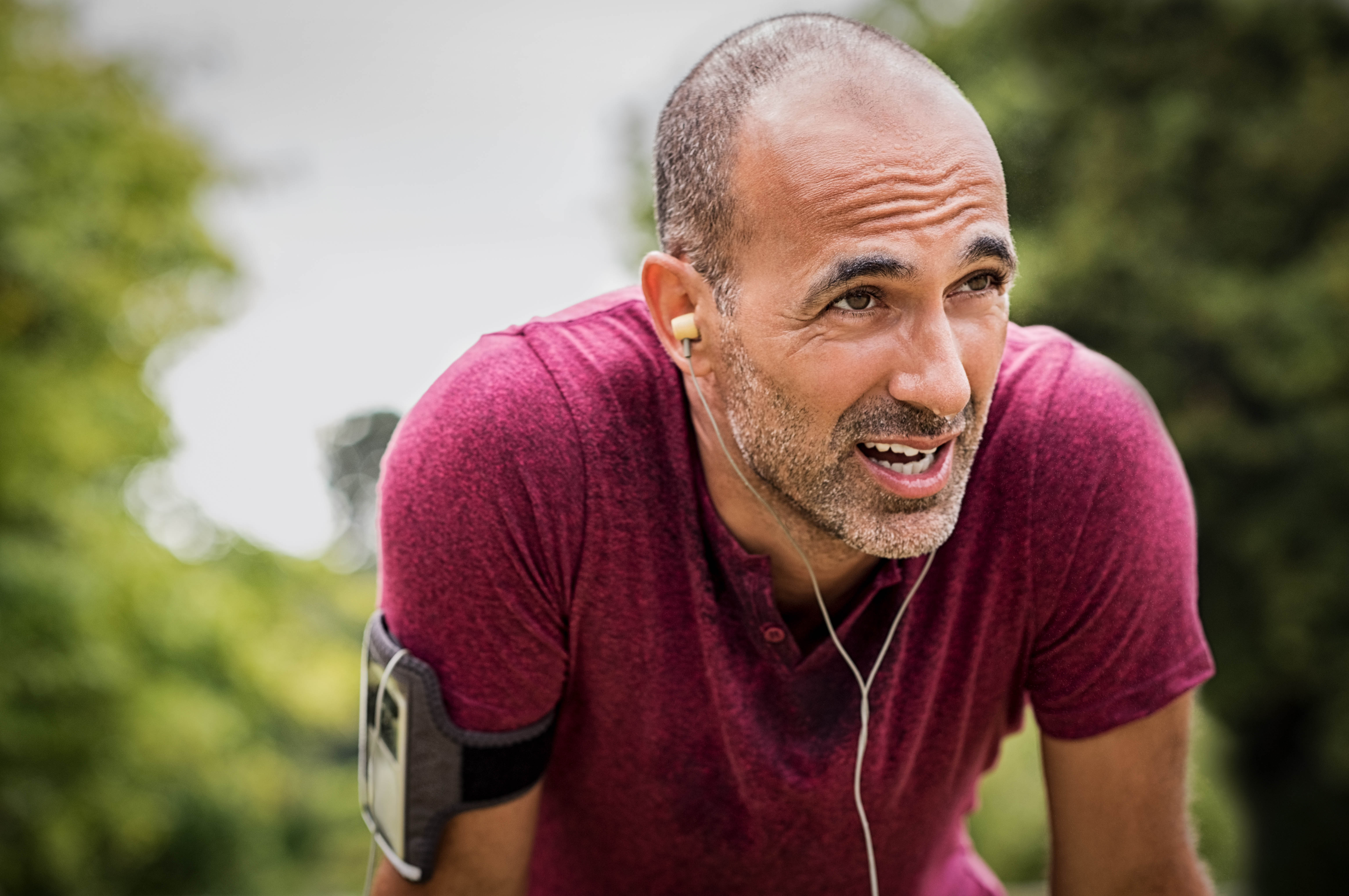 Middle aged man in a red t-shirt, hands on his knees, sweat dripping down his face, headphones in his ear, phone in a case on his arm Middle aged man in a red t-shirt, hands on his knees, sweat dripping down his face, headphones in his ear, phone in a case on his arm